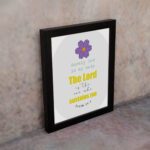 Psalm 54:4 Bible verse wall art with bold yellow text, purple flower, and light grey circular background pattern. displayed on the wall in a black frame