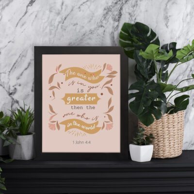 Infidu The One Who Is In You Bible Wall Art featuring 1 John 4:4 in golden-brown text on a soft pink background with floral and leaf patterns. displayed on the table in a black frame