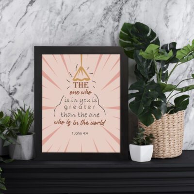 Infidu The One Who Is In You Bible Wall Art featuring 1 John 4:4 with brown and black text, light pink sunray background, and a lamp illustration. displayed on the table in a black frame