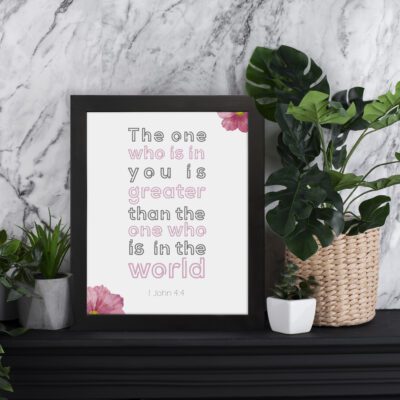 Infidu The One Who Is In You Bible Wall Art featuring 1 John 4:4 with light gray and pink text, white background, and floral accents. displayed on the table in a black frame