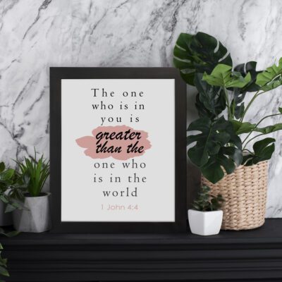 Infidu The One Who Is In You Bible Wall Art featuring 1 John 4:4 in bold black text with a pink brushstroke design. displayed on the table in a black frame