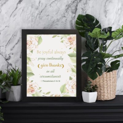 Infidu Be Joyful Always Bible Wall Art featuring 1 Thessalonians 5:16-18 with soft beige and green text and a floral design. displayed on the table in a black frame