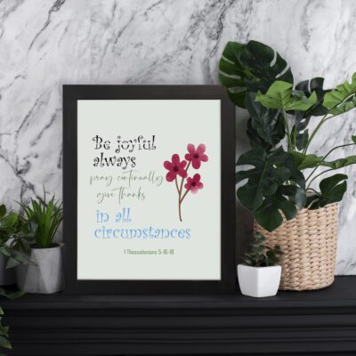 Christian wall art with the verse "Be Joyful Always, Pray Continually, Give Thanks in All Circumstances," with pink floral accents. displayed on the table in a black frame