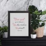 Psalm 115:3 Bible verse wall art with pink cursive and black text, floral accents, and a white background for a modern, spiritual design. displayed on the table in a black frame