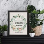 Rejoice always, pray without ceasing, give thanks in all circumstances 1 Thessalonians 5:16-17 Bible Wall Art with delicate floral wreath in pastel colors, light cream background in black frame displayed on the table