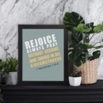 Rejoice always, pray without ceasing, give thanks in all circumstances 1 Thessalonians 5:16-17 Bible Wall Art with white and yellow text on muted teal background, bold design in black frame displayed on the table