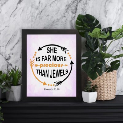 Infidu She Is Far More Precious Than Jewels Proverbs 31:10 Bible Wall Art with soft pink gradient, bold black text, and golden accents. displayed on the table in a black frame