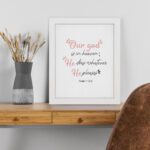 Psalm 115:3 Bible verse wall art with pink cursive and black text, floral accents, and a white background for a modern, spiritual design. displayed on the table in a white frame