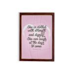 Proverbs 31:25 Christian wall art with She is clothed with strength and dignity on a soft pink background in an elegant script, framed. hung on the wall in a brown frame