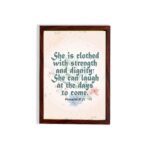 Wall art featuring Proverbs 31:25 with the quote in dark green text and floral decorations on a cream background. hung on the wall in a brown frame