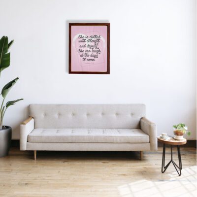 Proverbs 31:25 Christian wall art with She is clothed with strength and dignity on a soft pink background in an elegant script, framed. displayed on the wall in a brown frame