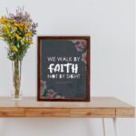 Christian wall art with 2 Corinthians 5:7 Bible verse on a light black background with a pink flower in the corner and white font and white framed on the table