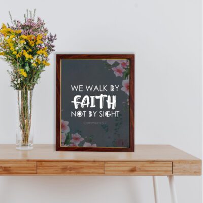 Christian wall art with 2 Corinthians 5:7 Bible verse on a light black background with a pink flower in the corner and white font and white framed on the table