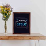 Bible wall art featuring the phrases Start With Jesus, Stay With Jesus, and End With Jesus in white and light blue text on a dark background. displayed on the table in a brown frame