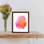 Bible wall art featuring Start With Jesus, Stay With Jesus and End With Jesus in white and orange fonts over a pink, orange, and yellow watercolor background. displayed on the table in a brown frame