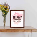 Bible wall art featuring The Lord Is My Rock, My Fortress, and My Deliverer in black handwritten-style lettering on a soft pink watercolor background. displayed on the table in a brown frame