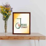 Jesus Christ text in green script with red floral accents on a glowing yellow-orange background, framed in black. displayed on the table in a brown frame