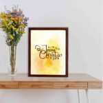 Jesus Christ Bible Wall Art in black script with a yellow cross and watercolor background, framed in black. Perfect Christian home decor. displayed on the table in a brown frame