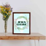 Infidu Jesus Christ Bible Wall Art with JESUS CHRIST text in a mint blue background surrounded by a floral wreath. displayed on the table in a brown frame