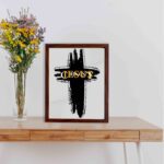 Infidu Jesus Christ Bible Wall Art featuring a black cross and gold lettering on a light gray background. Perfect for home decor. displayed on the table in a brown frame