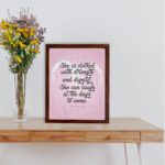 Proverbs 31:25 Christian wall art with She is clothed with strength and dignity on a soft pink background in an elegant script, framed. displayed on the table in a brown frame