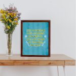 Christian wall art with Proverbs 31:25 verse in bold yellow text on a dark blue background with floral accents displayed on the table in a brown frame