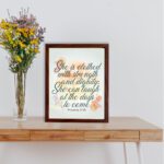 Christian wall art featuring the quote She is clothed with strength and dignity; she can laugh at the days to come from Proverbs 31:25, with an elegant design and pastel colors. displayed on the table in a brown frame