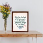 Wall art featuring Proverbs 31:25 with the quote in dark green text and floral decorations on a cream background. displayed on the table in a brown frame