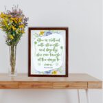 Christian wall art featuring Proverbs 31:25, with elegant script and floral accents in light green and gray background. displayed on the table in a brown frame