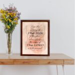 Bible wall art featuring Psalm 113:3 with floral elements and stylish text on a peach background. displayed on the table in a brown frame