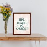 Christian wall art featuring the phrase His Grace Is Enough in teal lettering on a soft pink background with decorative swirls. displayed on the table in a brown frame