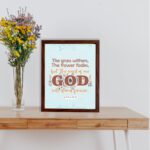 The Grass Withers Christian wall art with the verse Isaiah 40:8 in a modern font, featuring floral illustrations and a light blue background. displayed the table in a brown frame