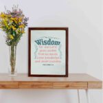 Wisdom Proverbs 2:6 Christian wall art with teal and red text on a mint green background in a black frame. displayed on the table in a brown frame