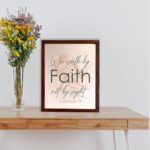 Christian wall art displaying the quote We Walk By Faith Not By Sight in elegant fonts on a soft beige-pink background. displayed on the table in a brown frame