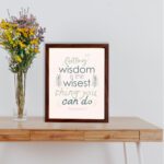 Infidu wall art with Proverbs 4:7 text surrounded by green leaves on a beige background. Elegant and minimal design. displayed on the table in a brown frame