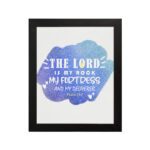 Bible wall art with The Lord Is My Rock, My Fortress, and My Deliverer in white stencil-style text on a blue watercolor background. hung on the wall in a black frame