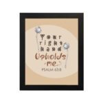 Christian wall art with the phrase "Your Right Hand Upholds Me" Psalm 63:8, featuring bold text and floral accents on a peach background. hung on the wall in a black frame