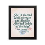 Wall art featuring Proverbs 31:25 with the quote in dark green text and floral decorations on a cream background. hung on the wall in a black frame