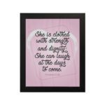 Proverbs 31:25 Christian wall art with She is clothed with strength and dignity on a soft pink background in an elegant script, framed. hung on the wall in a black frame