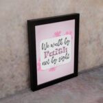 Christian wall art displaying We Walk By Faith Not By Sight with floral elements on a pale pink background. displayed on the wall in a black frame