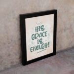 Christian wall art featuring the phrase His Grace Is Enough in teal lettering on a soft pink background with decorative swirls. displayed on the wall in a black frame