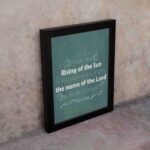 Wall art displaying Psalm 113:3, featuring white text on a dark green background with butterfly and leaf designs. displayed on the wall in a black frame