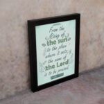 Wall art displaying Psalm 113:3 with elegant cursive font and leafy motifs on a mint green background. displayed on the wall in a black frame