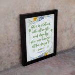 Christian wall art featuring Proverbs 31:25, with elegant script and floral accents in light green and gray background. displayed on the wall in a black frame