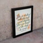 Christian wall art featuring the quote She is clothed with strength and dignity; she can laugh at the days to come from Proverbs 31:25, with an elegant design and pastel colors. displayed on the wall in a black frame