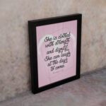 Proverbs 31:25 Christian wall art with She is clothed with strength and dignity on a soft pink background in an elegant script, framed. displayed on the wall in a black frame