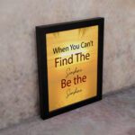 Infidu When You Can't Find The Sunshine Be The Sunshine wall art with a yellow background brown text and black frame on the wall. perfect for inspiring positivity in any workspace.