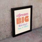 Infidu Dream Big Work Hard Make It Happen wall art with a light pink background and vibrant pink, orange, violet, and sky blue text and a black frame on the wall. Inspiring decor for home offices and workspaces.