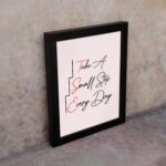 Infidu Take A Small Step Everyday wall art with a gray and light pink background and black and red text and a black frame on the wall. Inspiring decor for any workspace.