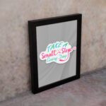 Infidu Take A Small Step Everyday wall art with a gray background and vibrant green, yellow, and pink text and black frame on the wall. Inspiring decor for any workspace.